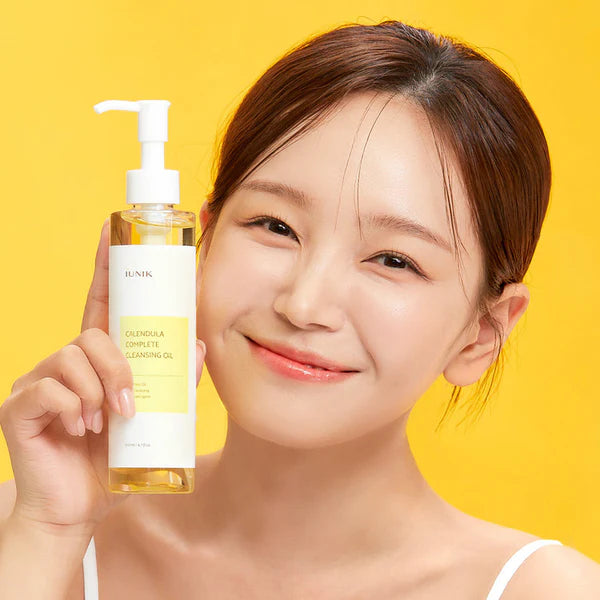 Smiling woman holding the iUNIK Calendula Complete Cleansing Oil bottle on a yellow background, Korean cleanser in Jeddah