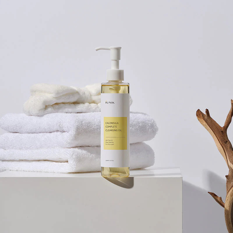 iUNIK Calendula Complete Cleansing Oil bottle styled with folded white towels and a dried branch