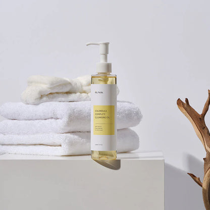 iUNIK Calendula Complete Cleansing Oil bottle styled with folded white towels and a dried branch