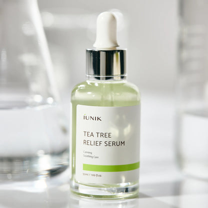 iUNIK Tea Tree Relief Serum dropper bottle standing among glass props in a bright, clean setting